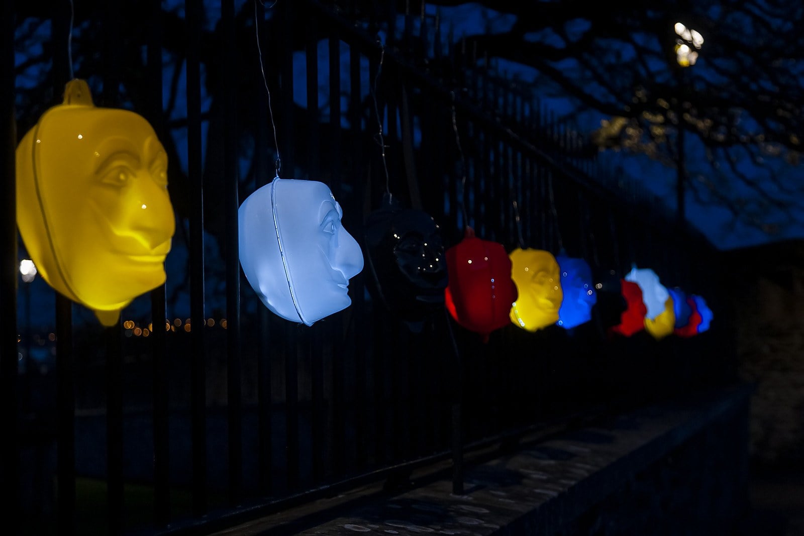Lit masks in yellow, white, red and blue hang along a railing on Derry's City Walls at dusk