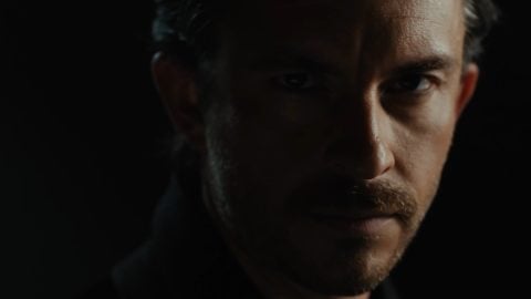 A man with dark hair and a mustache looks out from deep shadow in a still from the trailer for Audible's Dracula