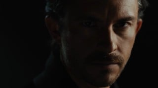A man with dark hair and a mustache looks out from deep shadow in a still from the trailer for Audible's Dracula