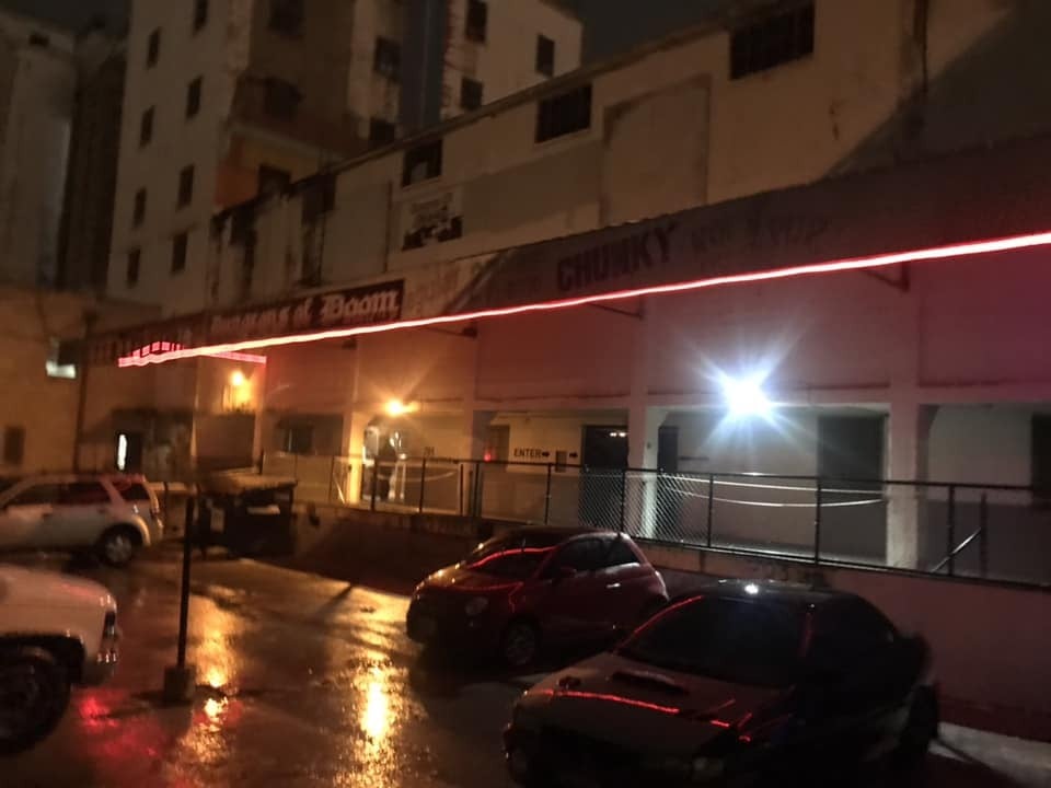 A long two-story building at night with a red neon strip along its awning and cars parked in front