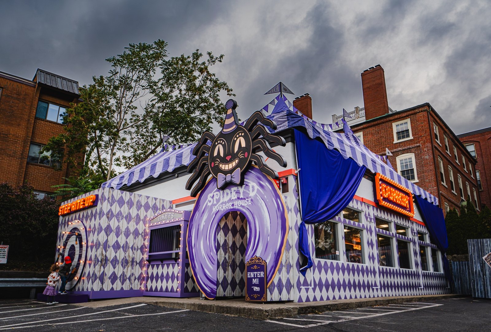 A restaurant wrapped in purple and white diamond patterns with a striped tent roof and a giant cartoon spider in a party hat above a donut-shaped entrance