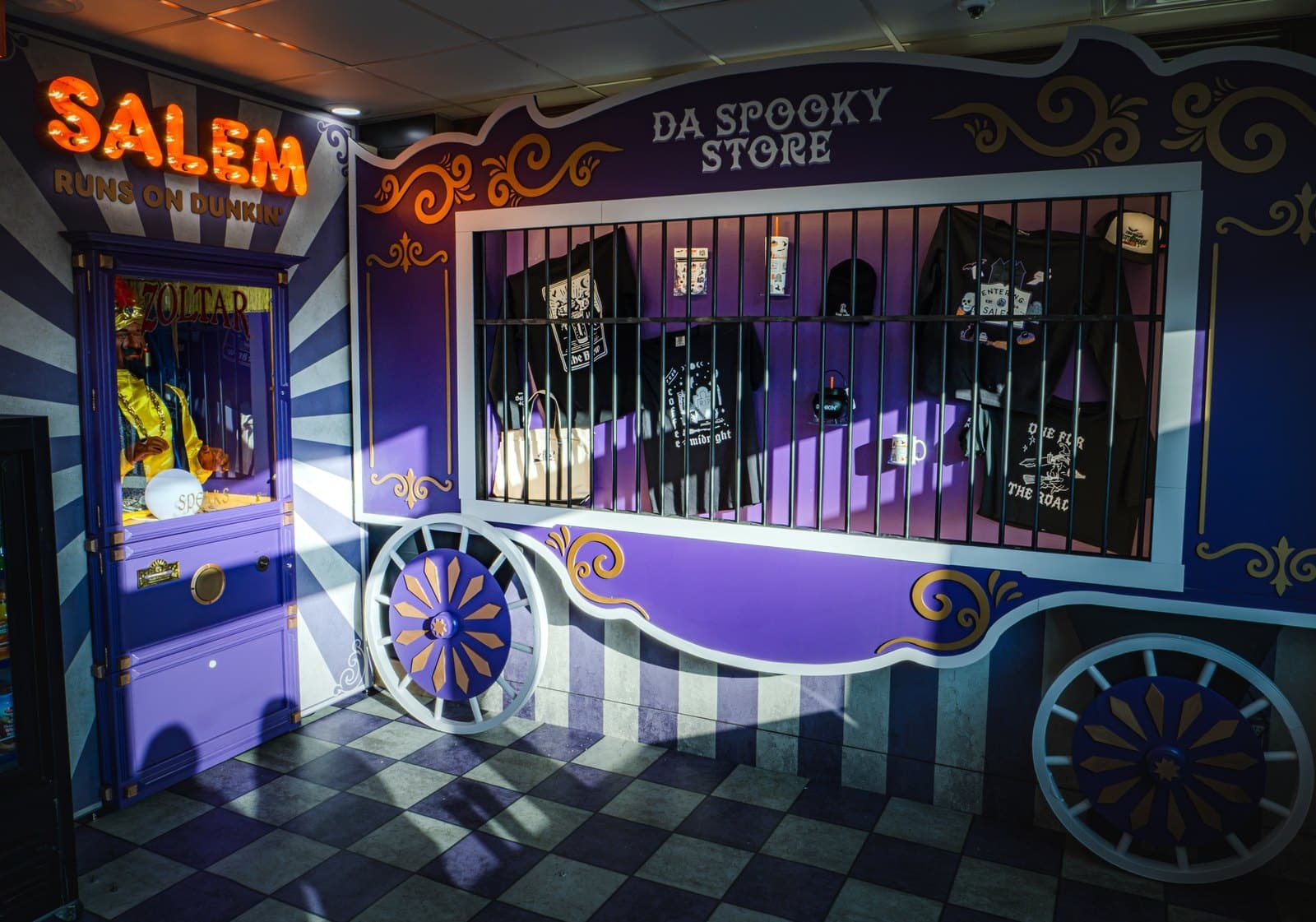 A purple circus wagon display with a barred window holding T-shirts and tote bags, beside a fortune teller machine, on a checkered floor