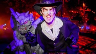 A performer in a wide-brimmed hat and ruffled shirt bares fangs beside a purple gargoyle statue at night