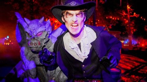 A performer in a wide-brimmed hat and ruffled shirt bares fangs beside a purple gargoyle statue at night