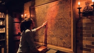 A woman points to a spot on a large antique city map on a brick wall, lit by candle sconces in a dark room