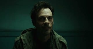 A man with dark tousled hair and scars on one cheek, in a green field jacket, sits in front of a teal wall