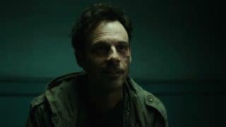 A man with dark tousled hair and scars on one cheek, in a green field jacket, sits in front of a teal wall