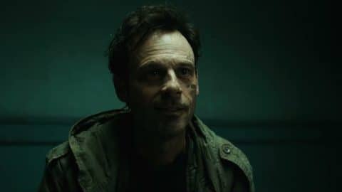 A man with dark tousled hair and scars on one cheek, in a green field jacket, sits in front of a teal wall
