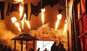 Flames shoot into the night sky over a crowd at FEAR Scream Park, with laundry lines and a stage behind