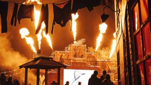 Flames shoot into the night sky over a crowd at FEAR Scream Park, with laundry lines and a stage behind