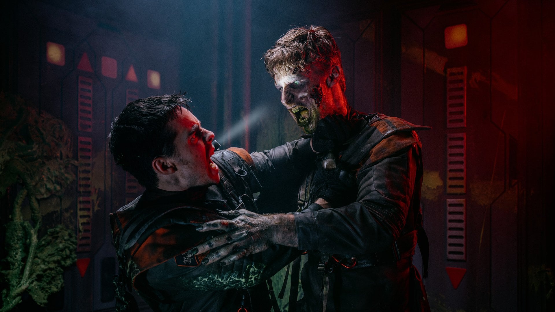 A performer in a dark flight suit with pale infected skin grabs another performer by the collar in a dim red-lit corridor