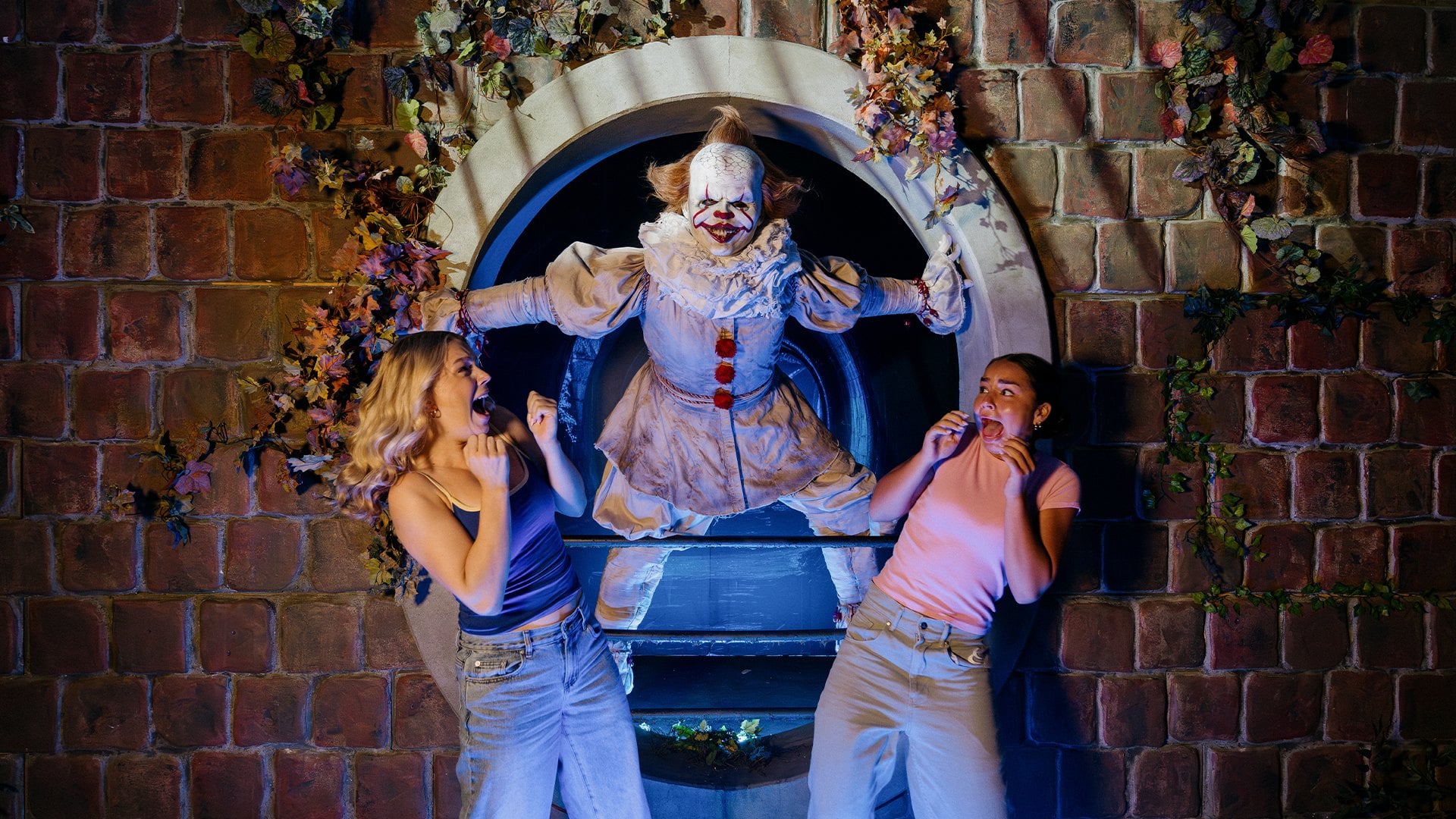 A performer dressed as Pennywise leans out of a round sewer opening in a brick wall between two guests who are reacting