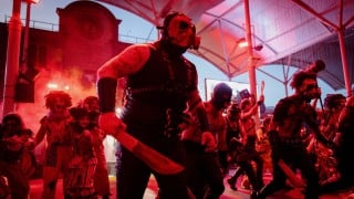 Costumed performers in masks and clown makeup charge down a park street in red light and smoke, led by a figure in a gas mask holding a prop machete