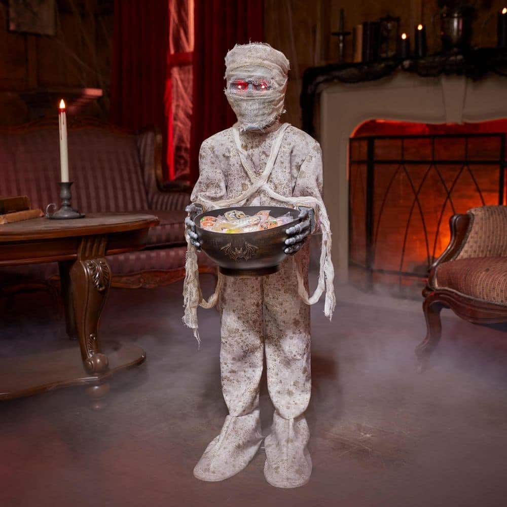 Gemmy 3 ft Animated Mummy with Candy Bowl at Home Depot: a child-height mummy in tattered gauze with glowing red eyes holding a black candy bowl in a dim room