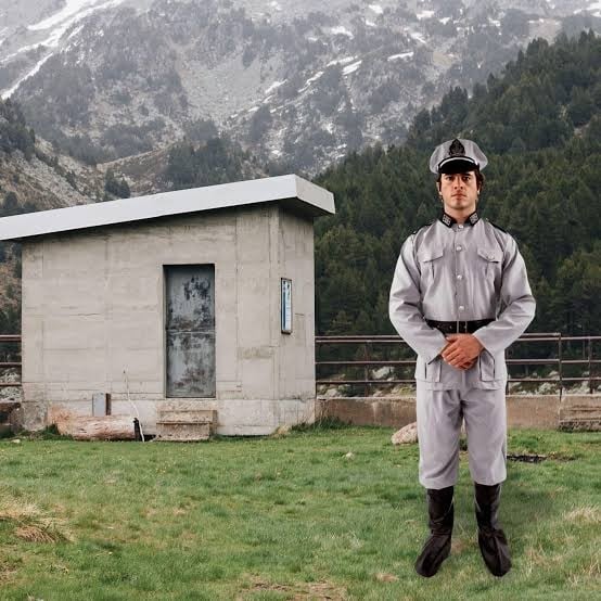 Second product listing image: the model in the gray German Army Soldier costume with boot covers, composited in front of a concrete hut and snow-capped mountains