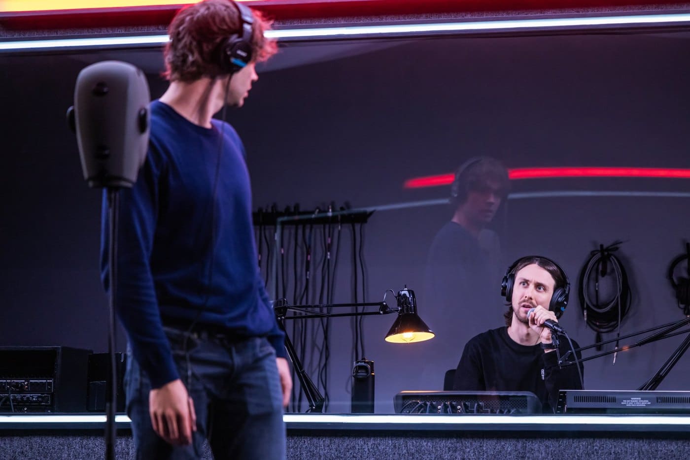 An actor in headphones looks back toward a second actor speaking into a microphone behind the glass of a recording booth