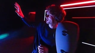 An actor in headphones raises a hand in red light beside a gray binaural microphone head in A Ghost in Your Ear