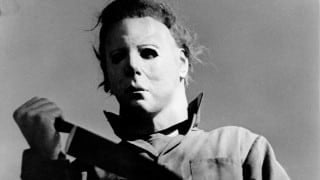 Black-and-white photo of a figure in a pale mask and coveralls holding a large kitchen knife, seen from below against a gray sky