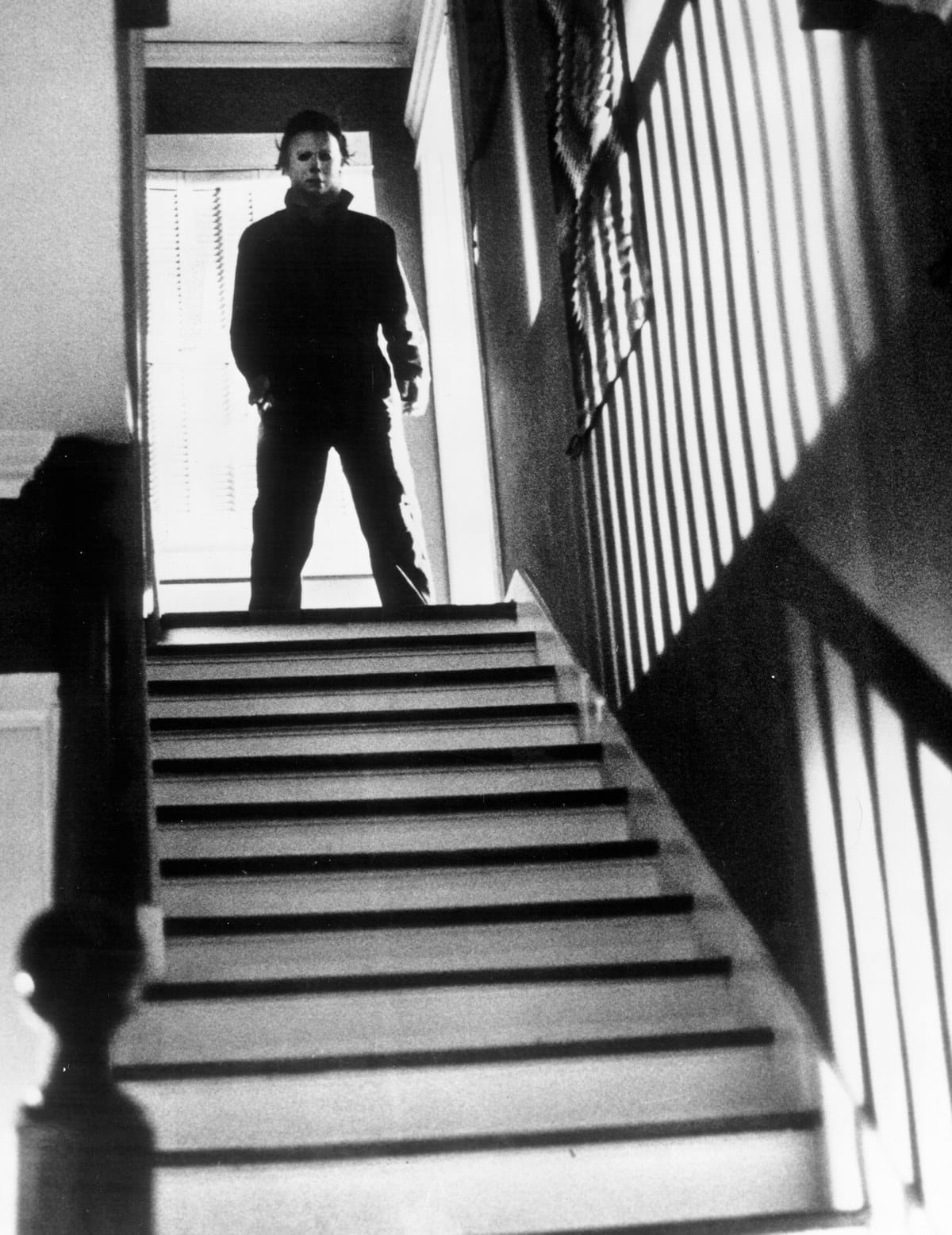 Black-and-white photo of a masked figure standing at the top of a staircase, lit from a window behind