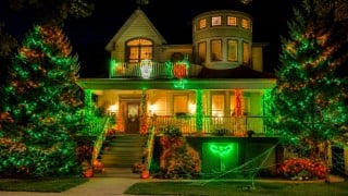 A two-story house at night outlined in green and orange lights, with lit trees on both sides, glowing pumpkin figures on the balcony and jack-o'-lanterns on the steps