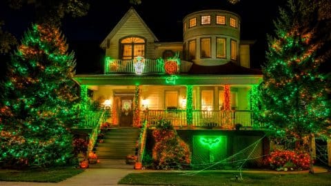 A two-story house at night outlined in green and orange lights, with lit trees on both sides, glowing pumpkin figures on the balcony and jack-o'-lanterns on the steps