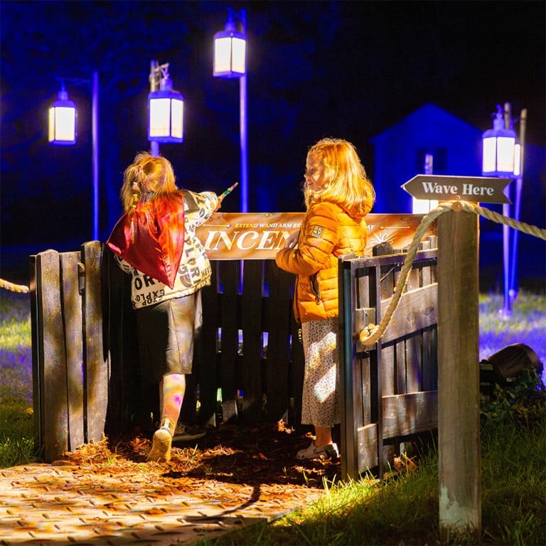 Two children lean over a wooden gate under lanterns in the lit village area at night