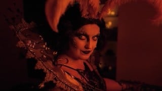 A performer with pale contact lenses, glitter eye makeup, and a red feathered headdress smirks under warm red light