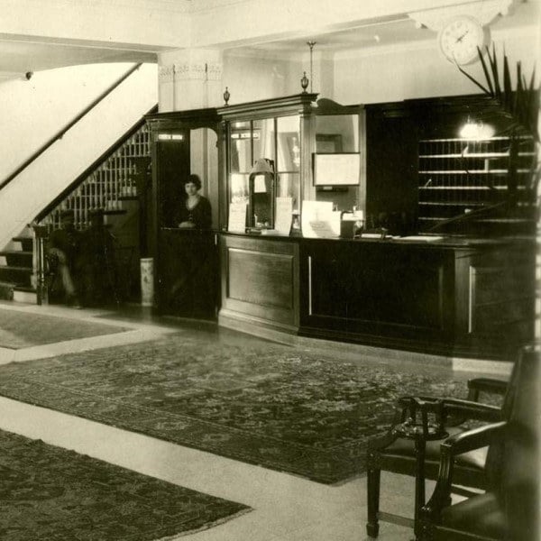 A sepia historical photo of a hotel lobby with a woman standing behind the front desk, a wall clock, and a staircase