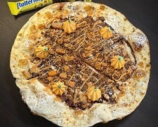 A round dessert pizza covered in chopped candy and a tan drizzle, with six piped orange pumpkins on top and powdered sugar on the crust, on a dark table