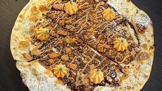 A round dessert pizza covered in chopped candy and a tan drizzle, with six piped orange pumpkins on top and powdered sugar on the crust, on a dark table