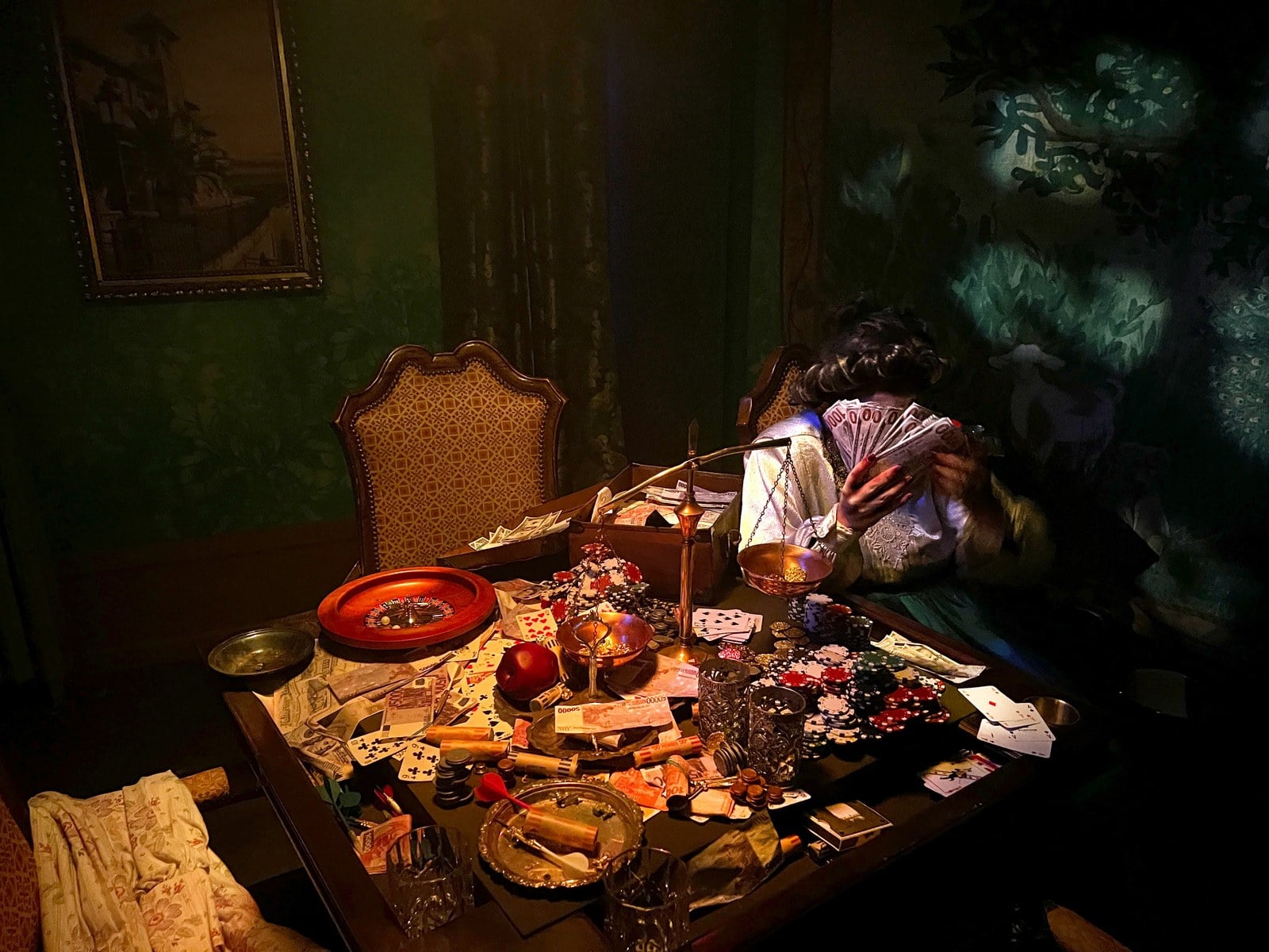 A card table heaped with chips, cards, cash, and a slumped figure in the Estate of Evil's greed room