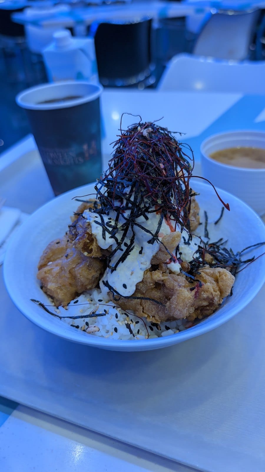 A bowl of fried chicken pieces and noodles with white sauce and a pile of dark shredded topping, on a blue-lit table