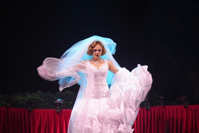 A performer in a white wedding dress swirls a long veil on a dark stage edged with skulls