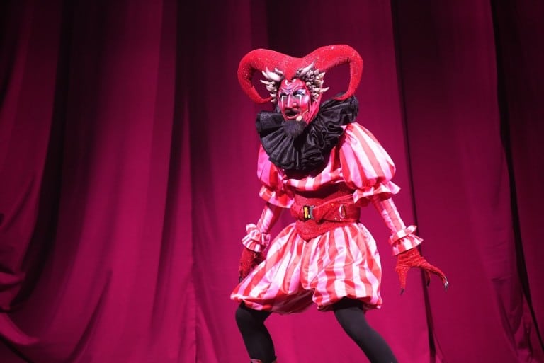 A performer in a red and white striped costume with red curled horns and a black ruff stands in front of a red curtain