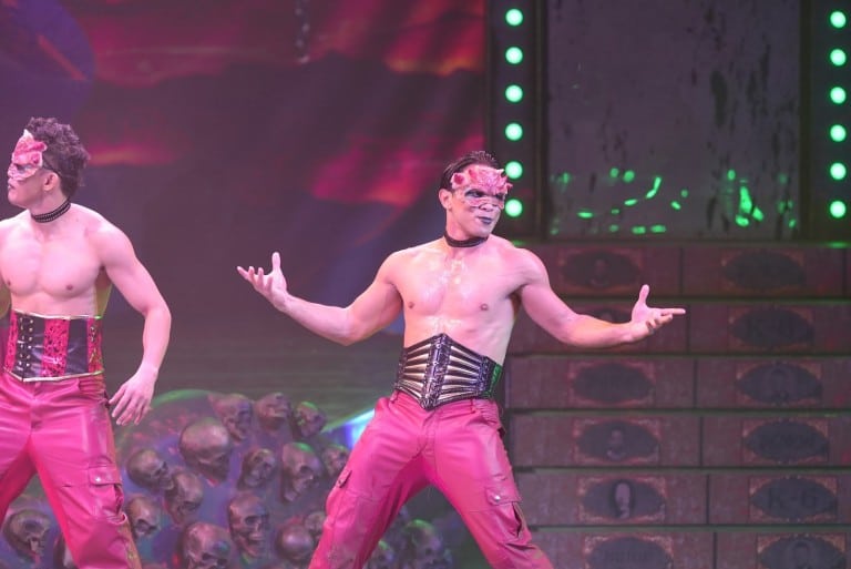 Two shirtless dancers in red masks, corsets and red trousers pose in front of a wall of skulls