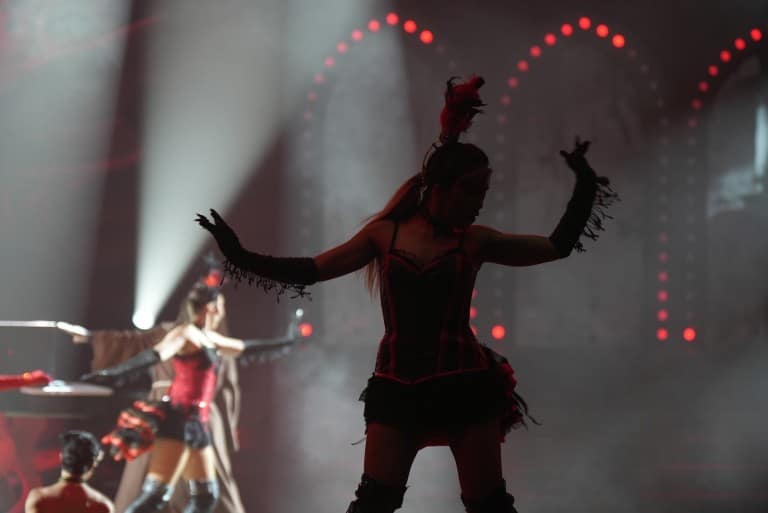 A dancer in a feathered headpiece stands in silhouette with arms out in white spotlights on a stage