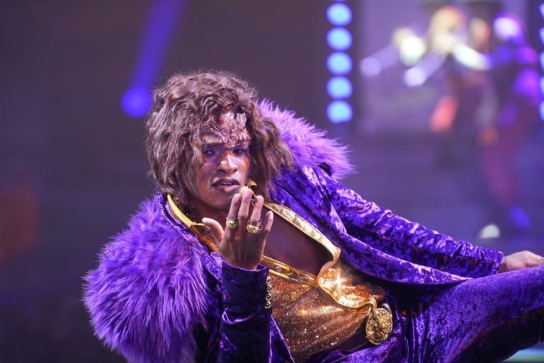 A performer in a purple fur-trimmed coat and gold shirt gestures toward the camera on a blue-lit stage
