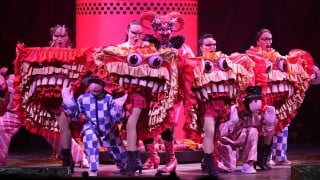 Dancers in red big-mouthed monster costumes fill a stage in front of a red cylinder set piece, with performers in harlequin checks in front