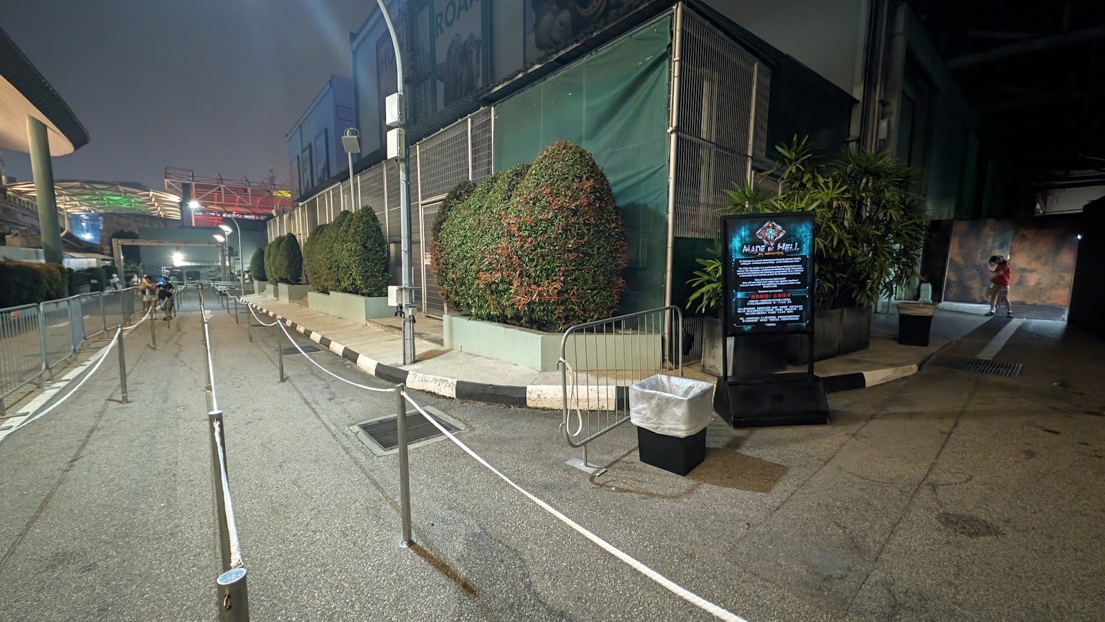 An empty outdoor queue lane with rope barriers beside a hedge and a lit sign, at night