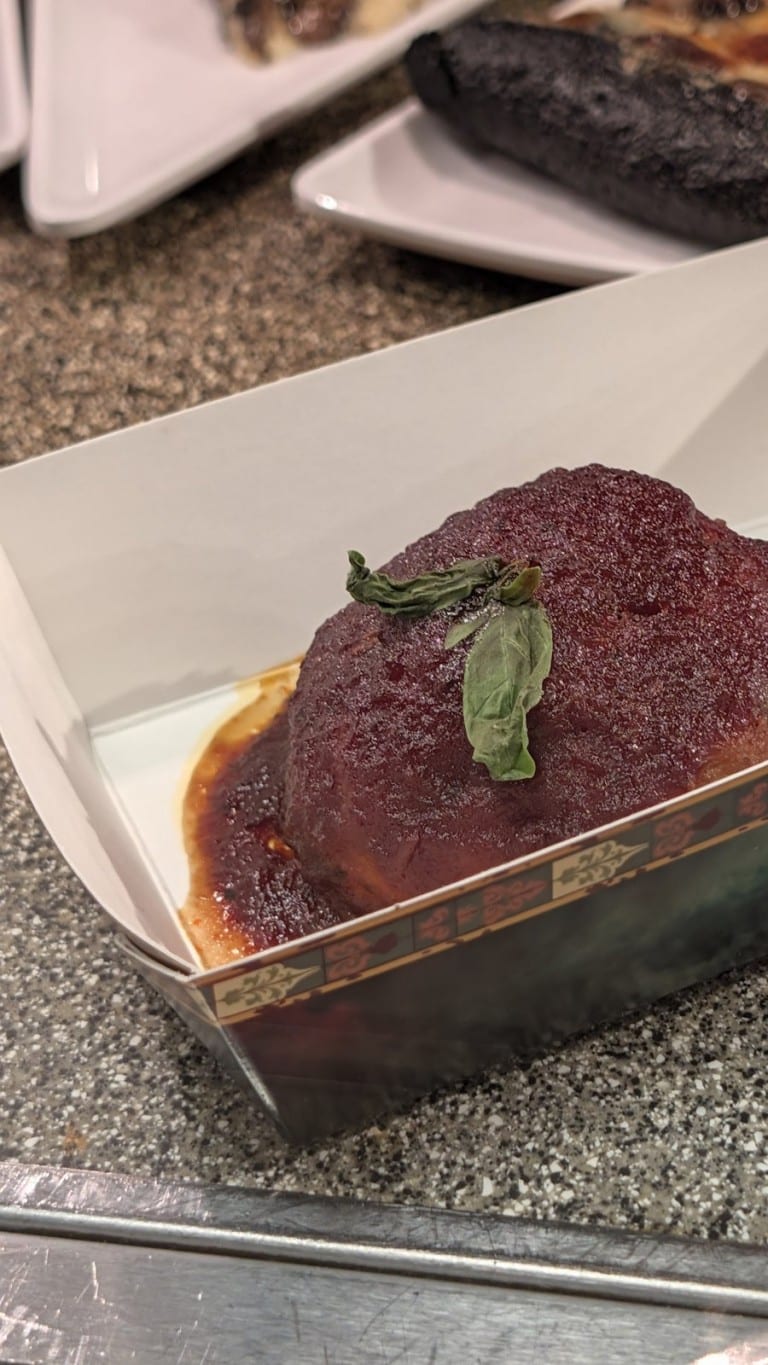 A large meatball in red sauce topped with a basil leaf in a paper tray