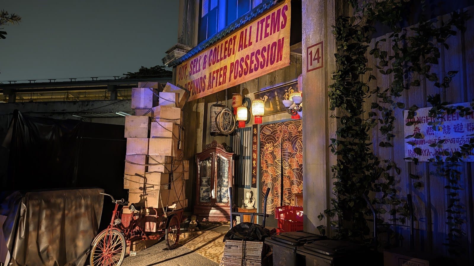 A cluttered shophouse front at night with stacked boxes, a bicycle, a red lantern and a banner across the top