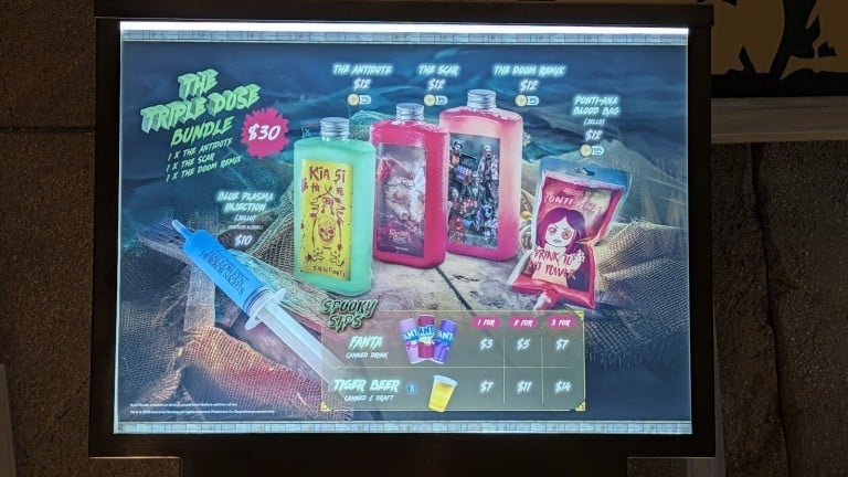 A lit drinks menu board picturing three flat bottles in green, red and pink, a red drink pouch and a blue syringe, with printed names and prices