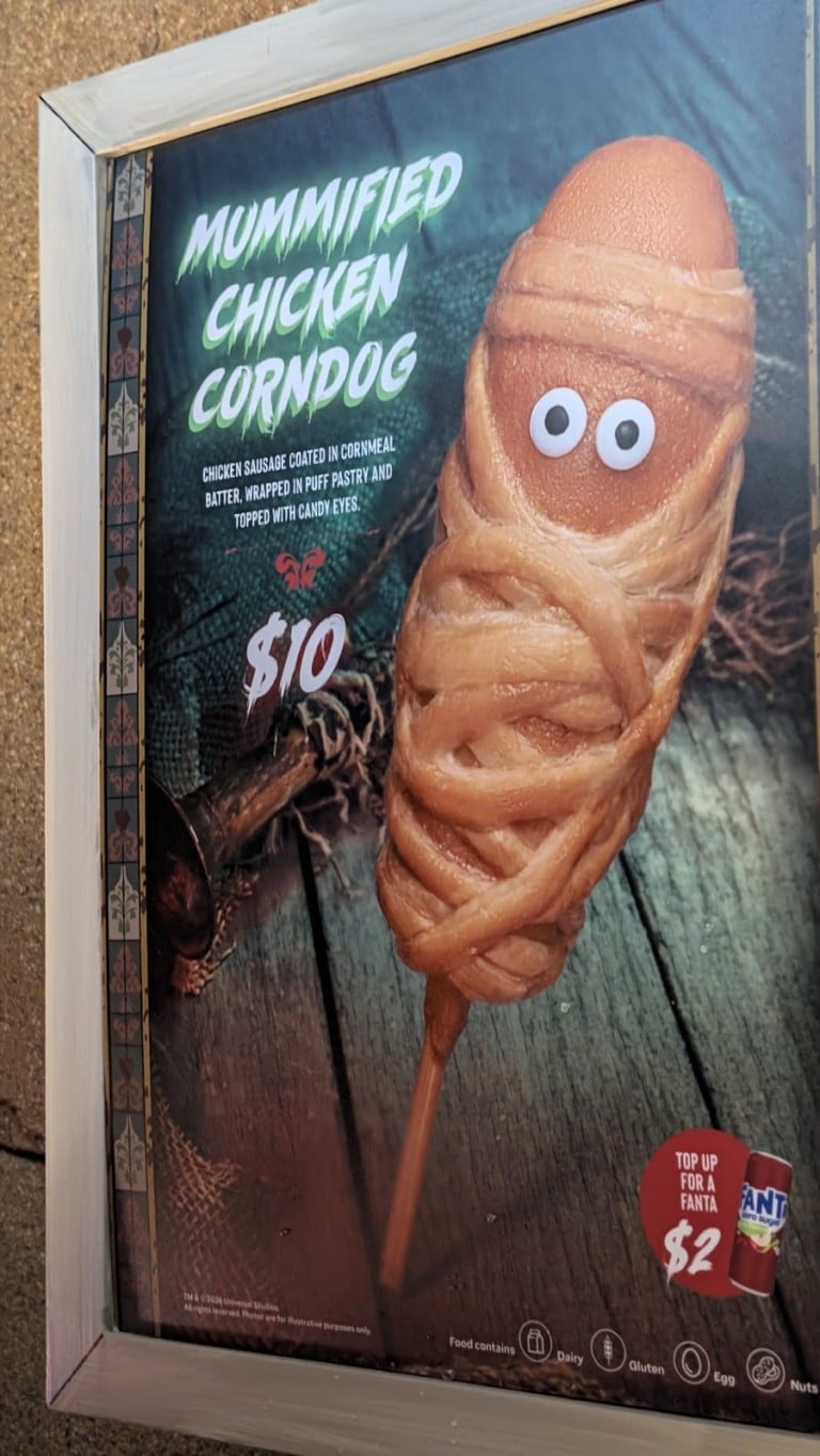 A framed menu sign picturing a corn dog wrapped in pastry strips with candy eyes and a printed name and price