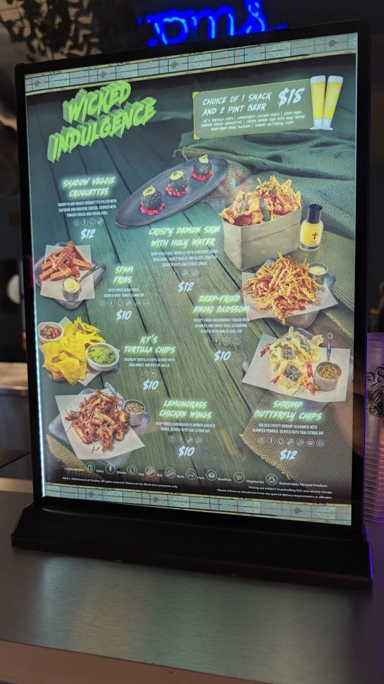 A lit menu board picturing seven fried snacks in trays and bowls, each with a printed name and price, and two glasses of beer