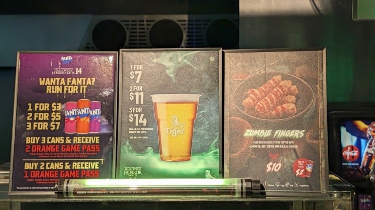 Three framed signs on a counter picturing soft drink cans, a cup of beer and a box of fried cheese sticks, with printed prices