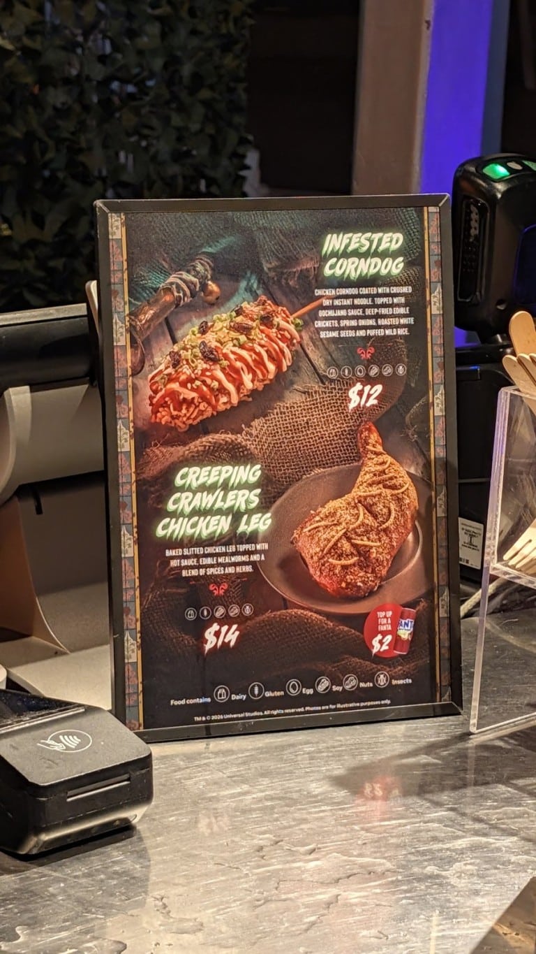 A framed menu sign picturing a corn dog and a chicken leg topped with insects, each with a printed name and price
