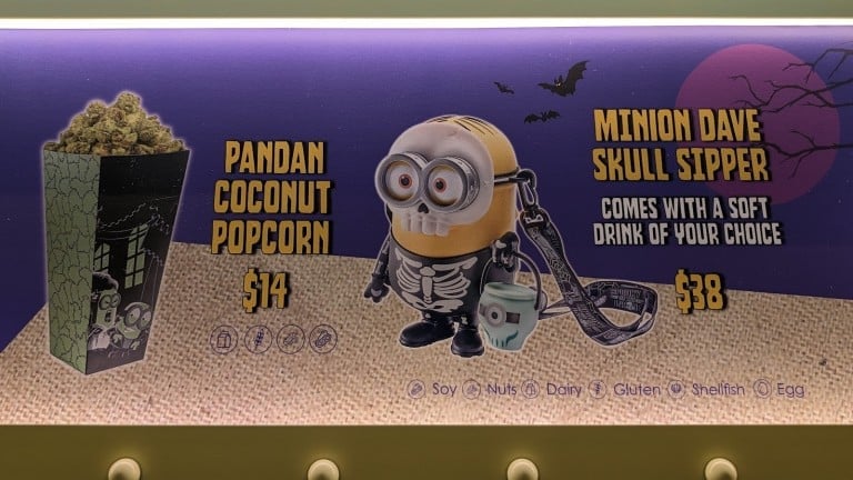 A purple menu sign picturing a popcorn box and a yellow cartoon character cup in a skeleton suit, each with a printed name and price