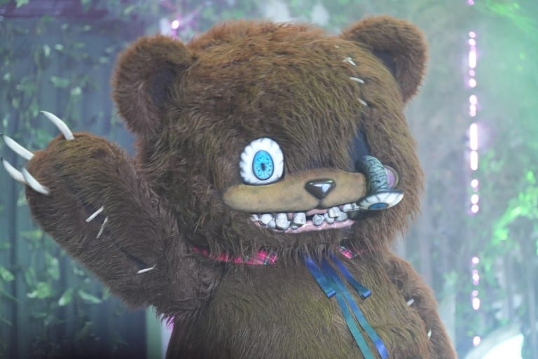 A brown teddy bear character with one blue eye, an eye patch, stitched teeth and long claws waves at the camera