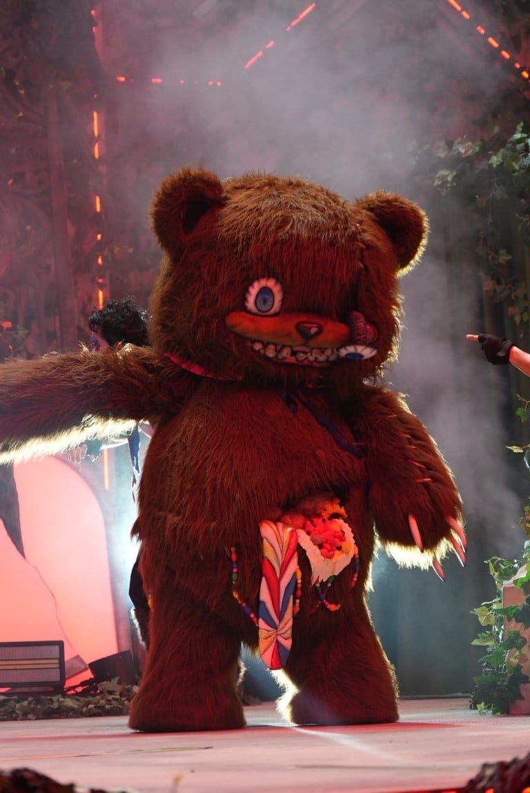 A brown teddy bear character stands on a foggy stage holding a striped bundle at its stomach