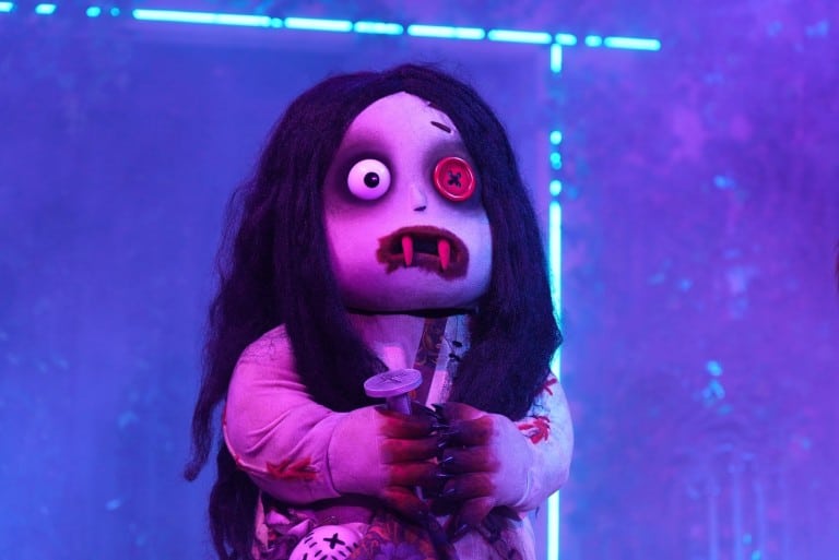 A costumed character with a large round head and one red eye stands in blue and purple light holding a small figure